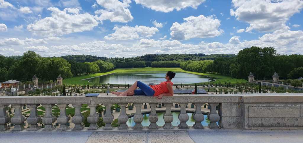 Woman with dark hair in bun when an orange shirt and long blue jean shorts leaning back on stone fence over looking the gardens of the palace of versailles with a lake and greenery in the background 