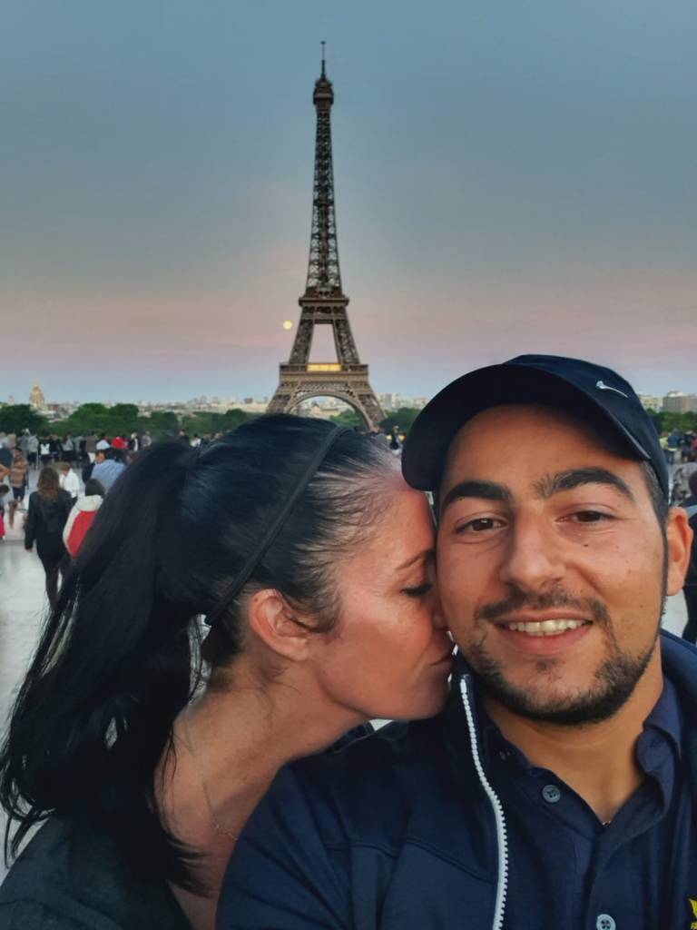 Couple with black haired ponytail woman with her nose nuzzled into smiling man wearing a black hats neck infront of the Eiffel Tower in Paris with sunset 