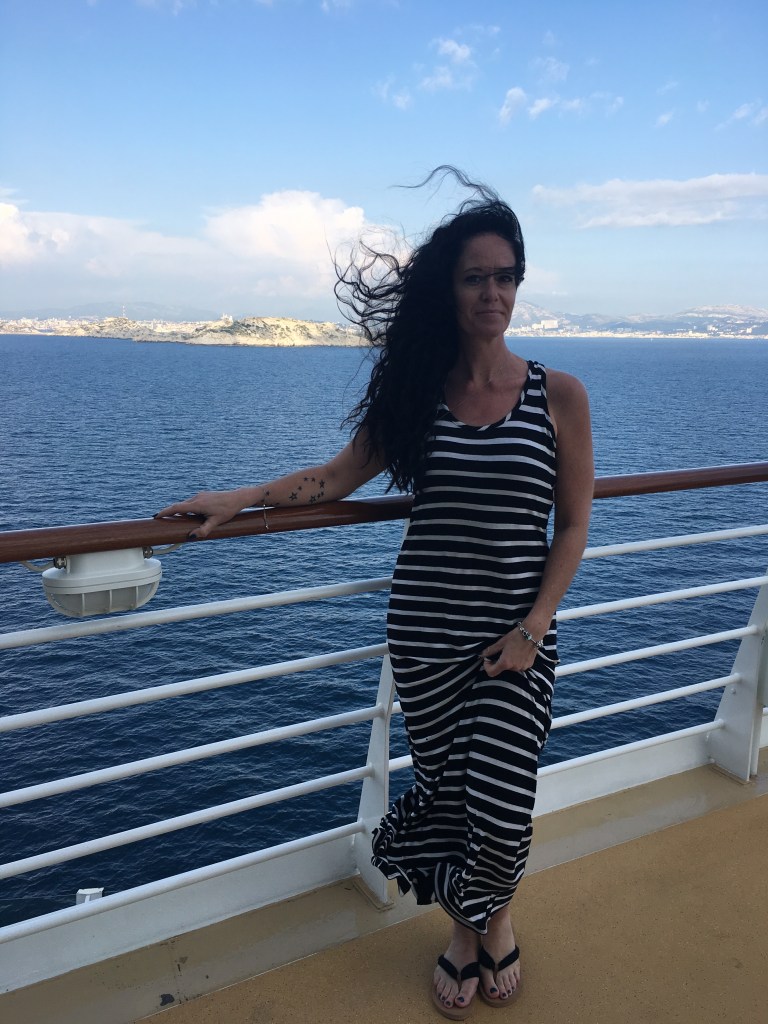 Woman with curly black hair with black and white striped summer dress smiling standing on the deck of the Royal Caribbean Symphony of the Seas Cruiseship with water in the background and landscape in the far distance