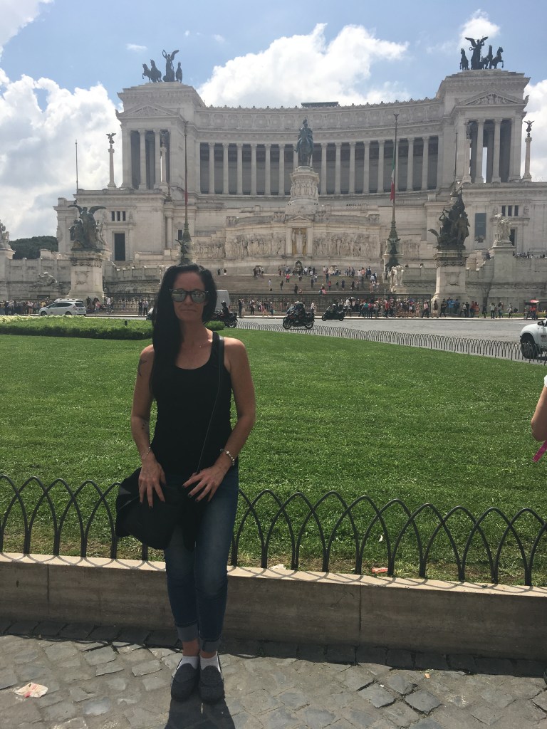 Woman with long black hair wearing blue jeans with a black tank top and holding a black purse standing in front of the Italian Parliment building with groomed green grass infront of the building