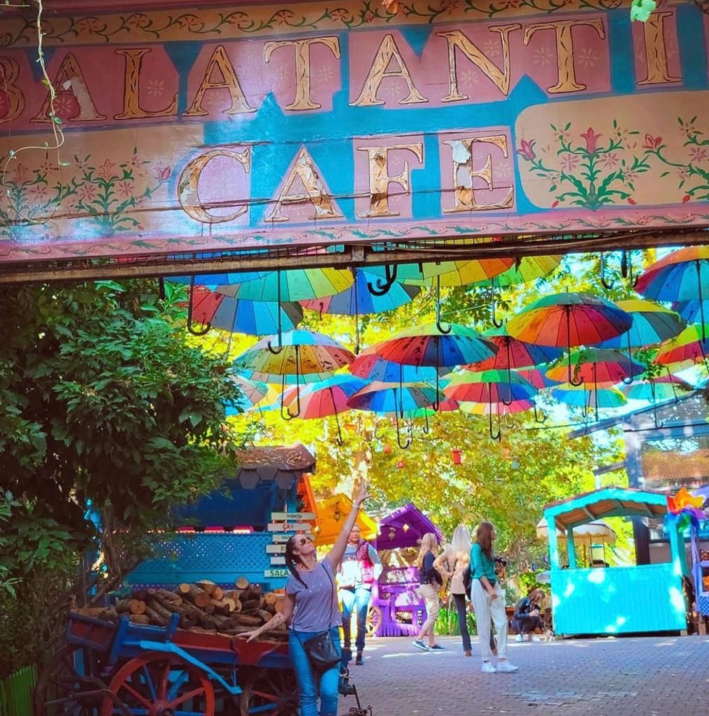 Balat Cafe sign in Istanbul with trees and beautiful colourful umbrellas lining up above with a woman reaching high up to touch them although they are out of reach as she leans on a blue old wheel barrel full of chopped wood