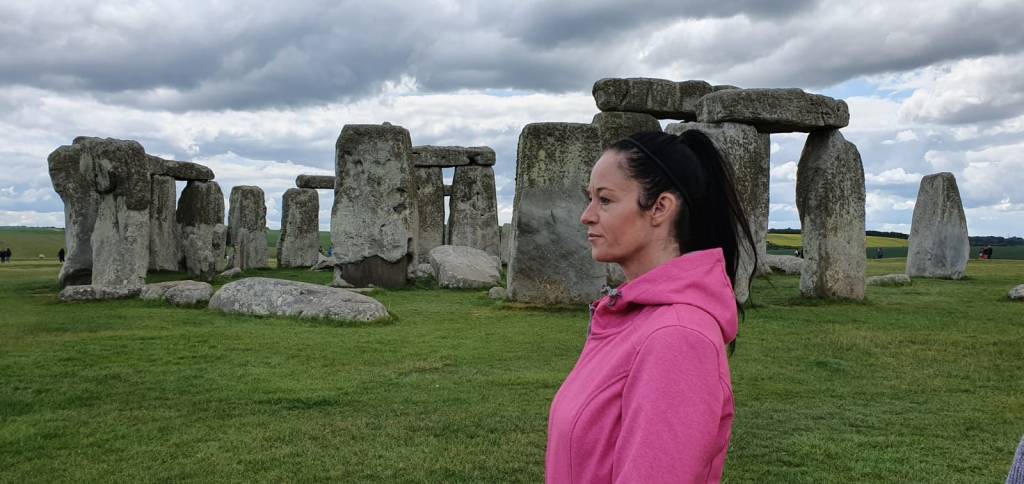 Stone Henge on a cloudy day with green grass and upper torso of woman with dark hair in ponytail with a pink coat posed standing in front of the Stone Henge
